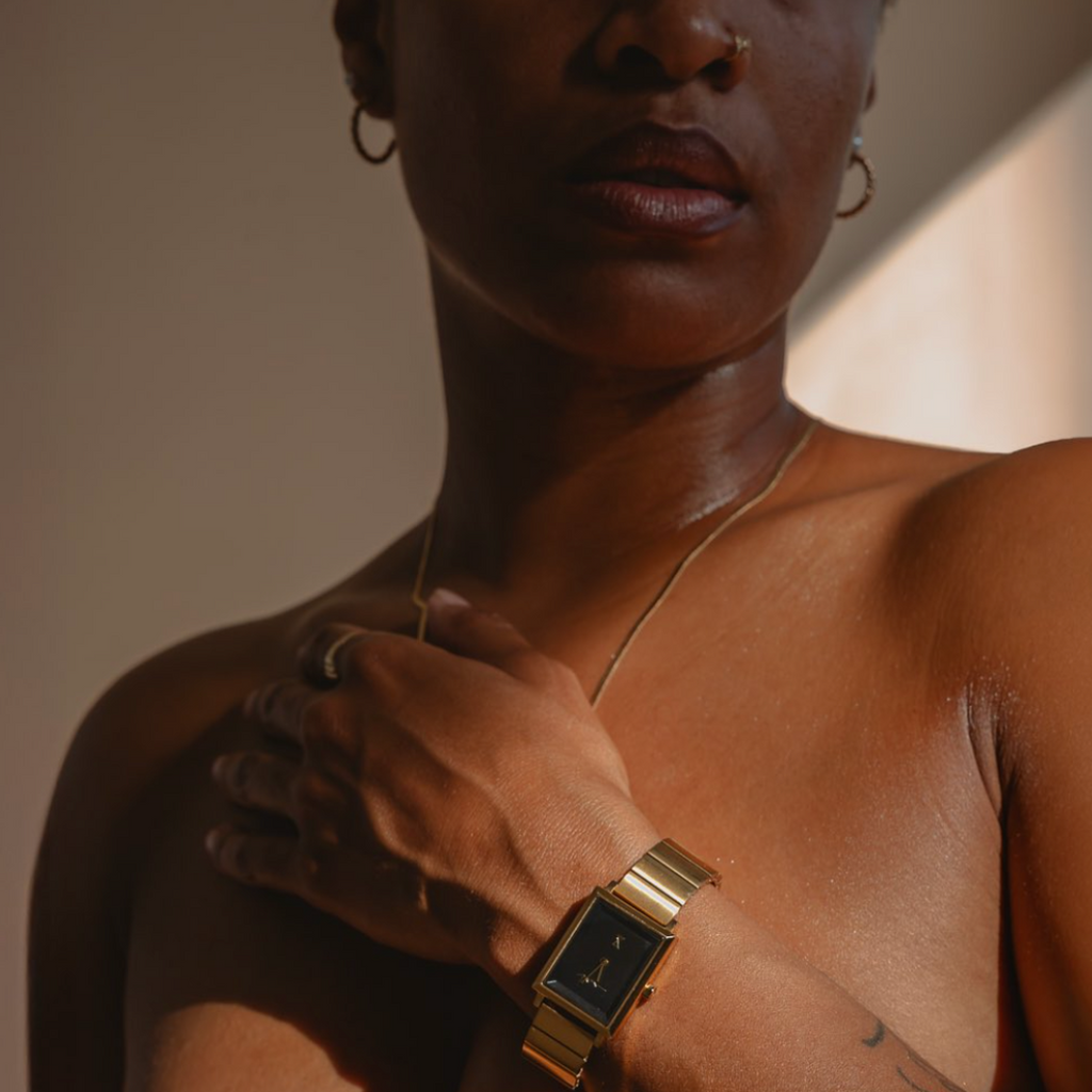 Close-up of a person wearing a gold watch and necklace against a neutral background